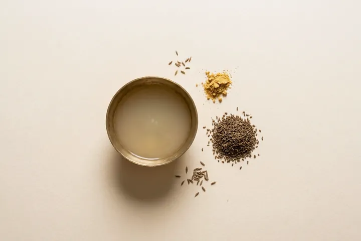 A brass bowl of cumin water with ajwain seeds and a pinch of hing beside it
